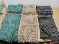 Six scarves laid out on a table: two green, two beige, two gray, all with dot texture and fringed edges.