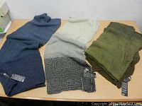 Photo of six scarves in shades of blue, green, beige gradient, and brown, all with 'MEN' tags laid out on a wooden surface.
