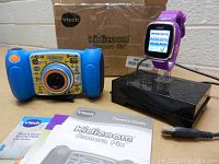 Photo showing blue Vtech Kidizoom Camera Pix with yellow front panel and orange shutter button along with purple Vtech Kidizoom smartwatch on black stand, charger, box, and manuals.