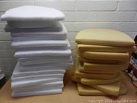 Two stacks of upholstery seat cushions shown, one stack white foam cushions and one stack yellow foam cushions on a table.