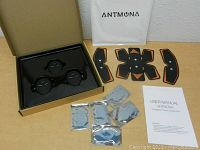 Overview of the open box showing three wireless control units, several adhesive electrode pads, user manual and branded pouch