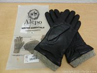 Pair of Alepo women's black sheepskin leather gloves with gray knit cuffs on a wooden surface next to the original Alepo packaging bag