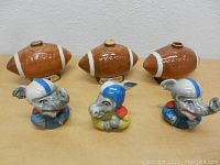 Six vintage ceramic Jim Beam whiskey decanters displayed on table, three shaped like brown footballs with white stripes and three shaped like cartoon animals wearing blue helmets.