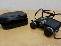 Vintage Promaster binoculars placed beside black zippered carrying case on a wooden surface.