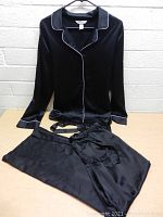 Black satin pajama set top and pants laid out on flat surface, showing button-up top with white piping detail and matching pants.