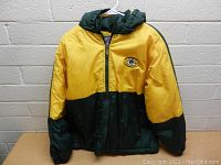 Front view of the Green Bay Packers boys size L jacket hanging on a white hanger against a concrete block wall, showing color blocking and logo detail.