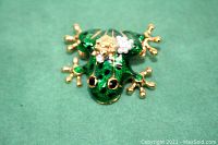 Front view of a green enameled frog brooch with black spots and gold-tone metal limbs, featuring a baby frog with pearl beads and rhinestone details on its back.