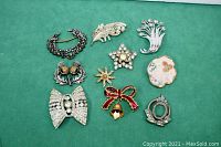 Full lot view showing all six vintage brooches arranged on green fabric. Variety of styles and materials visible, some embellishments missing or damaged.