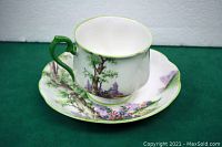 Side angled view of teacup and saucer showing green handle, countryside tree and floral decorations.