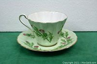 Teacup with handle and saucer showing the Scotch Thistle floral pattern on a green background with gold trim.