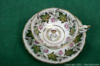 Full view of the teacup inside its matching saucer on a green background showing the floral and leaf pattern with gold trimming and handle accent.