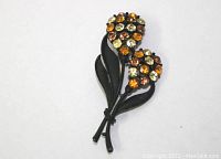Full view of the brooch showing the two flower heads, black stem and leaves, and multicolored rhinestones.