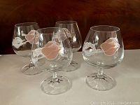 Four clear stemmed drinking glasses displayed on white surface showing floral decoration around the bowl.