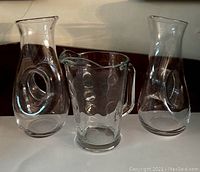 Front view of all three clear glass pitchers showing shape and design including the hollow circular handles on the two larger carafes and the dot pattern on the medium pitcher.