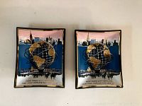 Pair of rectangular glass plates with Unisphere and New York City skyline design from the 1964-65 World's Fair. Plates show gold and black globe with orbit rings in front of a pink and black skyline with text.