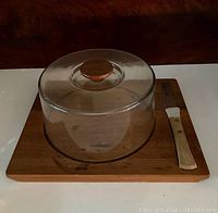 Side view of teak wood serving board with clear dome cover and cheese knife with light wood handle.