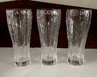 Three tall clear Iittala drinking glasses with textured bark-like vertical ridges and thick solid base shown side by side on dark background.
