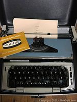 Front view of the blue Eaton Viking Deluxe 10 portable typewriter with black round keys, typed sheet sample loaded, black case visible underneath, and yellow owner's manual.