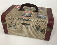 Full view of the vintage travel case showing front and top with metal clasps and brown handle; fabric exterior with country stickers