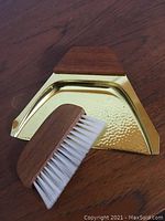 Brass crumb sweep tray with teak handle laid on wooden surface with the matching teak-handled brush with white bristles.