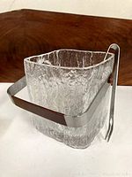 Front and side view of the vintage textured glass square ice bucket with metal handle and wooden grip, and ice tongs hanging on handle.