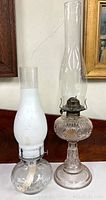 Lot of two glass oil lamps on wood surface, one with clear glass base and ornate pattern, the other with clear base and white frosted chimney