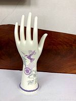 Off-white decorative hand display with purple bird flying and floral patterns mainly in purple, positioned upright on a white surface against a wood background.
