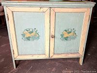 Front view of the vintage wooden cabinet showing two pale green doors with hand-painted fruit design and cream-colored framing.