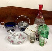 Nine decorative home items displayed on white surface against wood background, showing variety of porcelain, glass, and metal items including Limoges candle holder and Italian glassware.