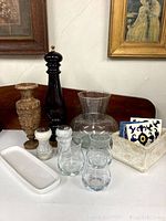 Photo showing carved wooden vase, tall dark wood pepper grinder, white marble salt and pepper shakers, clear glass vase/carafe, rectangular white ceramic tray, clear glass drinking glasses, stone-like container with ceramic tiles