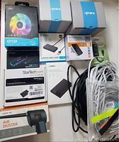 Photo showing boxed items including Deepcool CF120 fan, QGeeM USB-C hub, Orico USB hub, wireless N router, StarTech SSD adapter, Opolar air duster, and various cables.