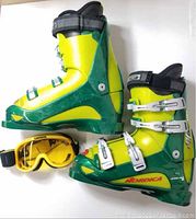 Side view of the pair of Nordica ski boots in green and yellow with four silver buckles and Carrera yellow goggles beside them.