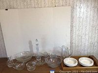 Vintage glassware and ceramic bowls arranged on a wooden surface against wallpapered wall. Includes glass pitchers, pressed glass bowls, crystal decanter with stopper, small glass bowls, and ceramic bowls on a tray.