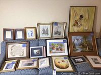 Overall group photo showing various framed paintings and prints arranged on a couch and wall, including inspirational needlework, vintage glass prints, woodland and 1970's boy and rabbit paintings.