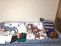 Wide shot of all textiles and decorations laid out on a blue couch, including afghans, linens, aprons, and plush items for overview of lot contents.