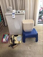 Wide shot showing white vintage hamper, off-white bin, blue stool, tray of small items, curling irons, mirror and scale on carpet beneath a window with curtains.