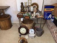 Wide view of various home decor items including two wall clocks on the floor, multiple baskets under and around tables, a chalkware figurine, framed prints, wooden birdhouse and other decorative pieces