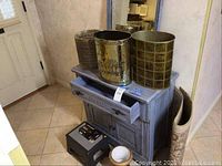 Photo of the blue wash stand with drawer partially open, three vintage metal waste cans on top, cream colored bowl on the floor, dark-colored step stool at bottom left, all against a wall with blue framed mirror behind.