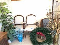 Overall view showing two beige carved armchairs, caned blue print side chair, artificial Philodendron, glass vase with blue dried flowers.