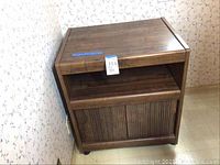 Brown veneer tea cart placed against a patterned wallpapered wall. Shows front and side with closed tambour-style cabinet door on lower section.