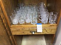 Photo showing the full lot of 31 glasses arranged inside a wooden cabinet drawer. Variety of glass types visible: water glasses and larger mugs with etched designs.