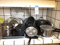 Wide view of assorted pots and pans on stove including egg poacher, enamel baker, pressure cooker, and others