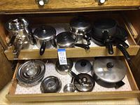 Cookware in two pull-out drawers showing pots, pans, lids, and a multi-tiered double boiler, all in worn condition.