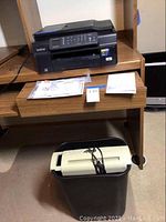 Portable paper shredder with black bin and white top positioned below the desk with power cord wrapped on top.