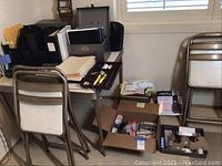 Photo showing folding chairs, table with papers and office supplies on it, and boxes containing various office items.