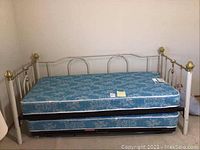 White metal trundle day bed with gold-tone finials, main mattress on top and trundle mattress pulled out below, showing floral blue patterned covers