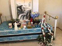 Photo of vintage Asian scroll painting, assorted candles, silk flowers in vase, macrame with silk plant, silver plated candle holder and faux crocodile leather jewelry holder on a blue patterned mattress