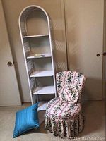White wicker five-shelf etagere next to white wicker upholstered chair with skirt, two pillows on floor in front