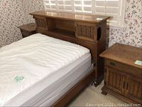 Wooden full bed with white mattress and two matching bedside tables against floral wallpaper, partial view of bed headboard with shelving and sliding doors