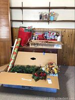 Overview photo showing boxed Christmas tree parts lying horizontally along the floor, several wreaths on the floor and on top of the box, wrapping paper roll, and other holiday decorations on a wooden cabinet surface in the background.